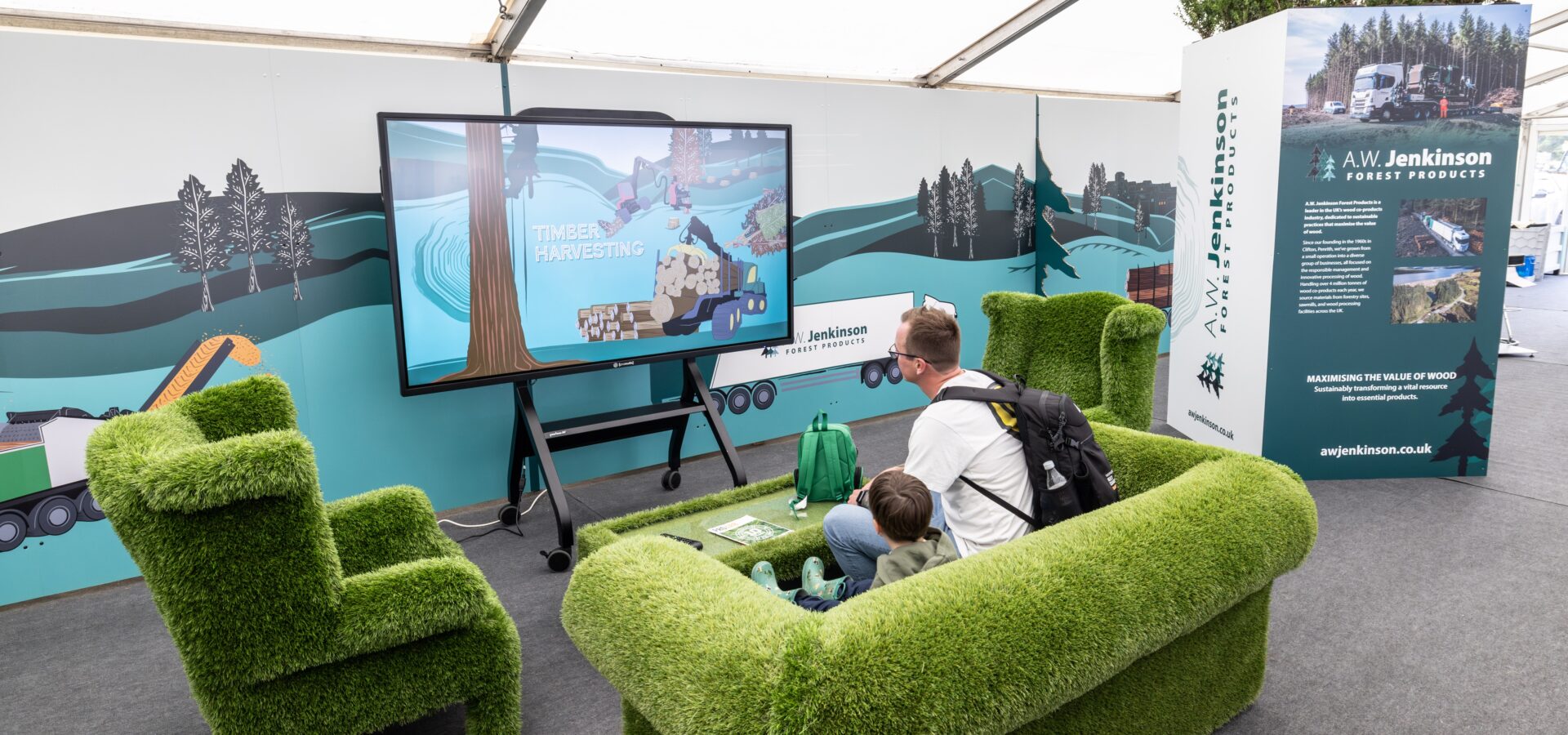 A man and child sit on the green sofas, watching an AW Jenkinson video on a TV