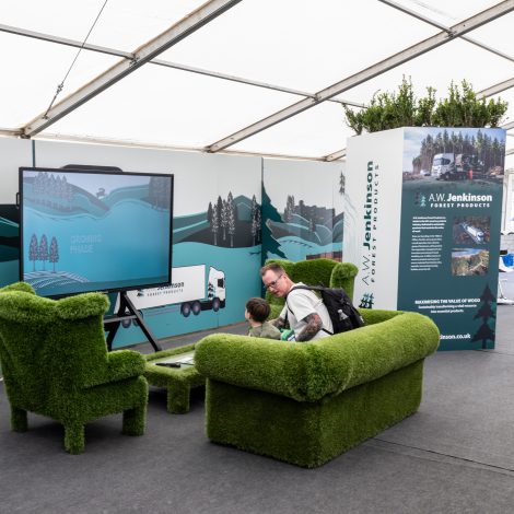 A man and child sit on the green sofas, watching an AW Jenkinson video on a TV