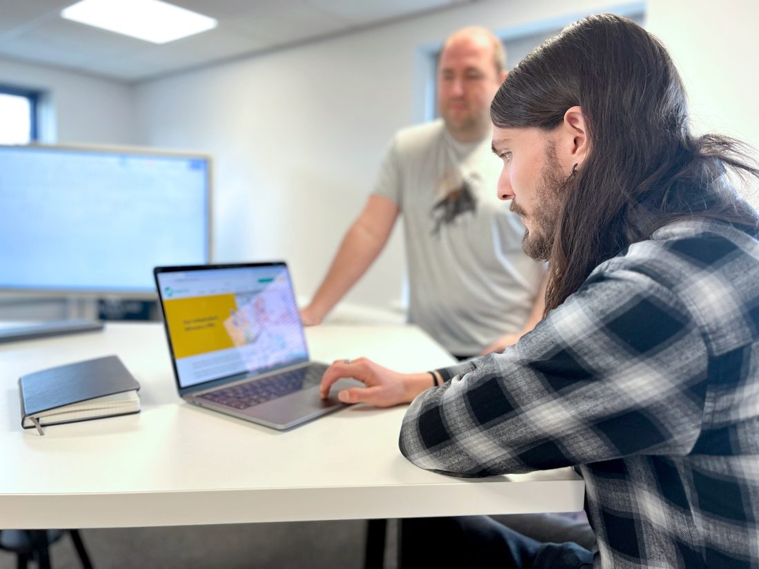 Luke working on a laptop at a desk with Chris standing leaning on the desk in the background.